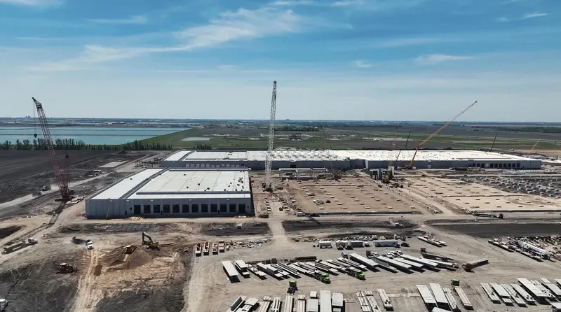 Harwood campus mid-build: precast wall panels erected and roofs decked on two buildings, crawler cranes working the line, racked precast panels in the laydown yard